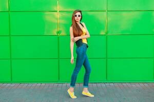 jovem lindo menina dentro oculos de sol dentro uma amarelo camiseta, jeans e tênis posando perto verde parede foto
