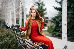 garota com cabelo vermelho em um vestido vermelho brilhante em um banco de um parque vazio foto