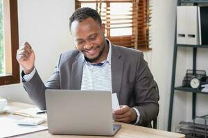 africano americano Preto homem feliz enquanto lendo companhia o negócio resultados em Smartphone e computador portátil. o negócio finança e tecnologia conceitos. foto