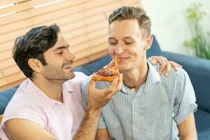 alegre gay casal comendo pizza. gay casal sentado em sofá às lar, comendo 1 fatia do pizza. homossexual relação conceito. foto