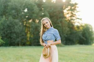 uma lindo jovem menina dentro luz roupas detém uma chapéu dentro dela mãos enquanto caminhando dentro uma compensação dentro a floresta foto