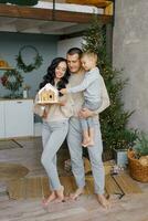 uma à moda jovem família com uma pequeno criança é em pé dentro seus cozinha, decorado para Natal, e admirando uma Pão de gengibre casa preparado com seus próprio mãos foto