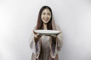 uma sorridente ásia muçulmano mulher é jejum e com fome e segurando e apontando para uma prato foto