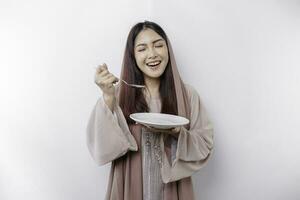 uma sorridente ásia muçulmano mulher é jejum e com fome e segurando e apontando para uma prato foto