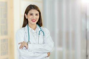 profissional jovem ásia mulher médico em pé com braços cruzado feliz e sorrir dentro exame quarto às hospital. vestindo uma branco roupão e estetoscópio. Novo normal e cuidados de saúde conceito. foto