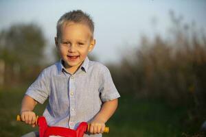 alegre pequeno Garoto é segurando uma vermelho leme do uma brinquedo motocicleta e sorridente para a Câmera. foto