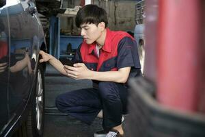 jovem masculino ásia profissional automotivo mecânico trabalhador Verificações pneu pressão de Smartphone inscrição às uma carro garagem, especialista dentro manutenção veículo serviço, e fixação ocupações auto indústria. foto