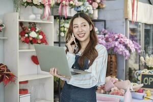 1 jovem ásia fêmea florista proprietário, trabalhando com computador portátil, vendendo floral arranjo, falando em Móvel telefone dentro colorida flor fazer compras loja com uma lindo grupo do flores, e comércio eletrônico negócios. foto