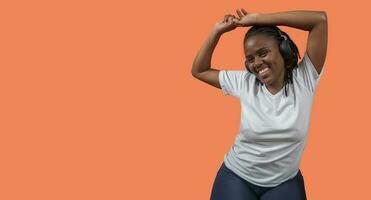 retrato do feliz jovem africano mulher dançando e ouvindo para música com fones de ouvido, braços acima, vestido dentro branco camiseta e azul calça contra laranja colori fundo foto