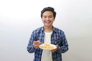 retrato do feliz ásia homem dentro azul xadrez camisa comendo delicioso instante Macarrão com pauzinhos servido em placa. isolado imagem em branco fundo foto