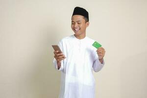 retrato do atraente ásia muçulmano homem dentro branco camisa com calota craniana segurando uma Móvel telefone e apresentando crédito cartão. isolado imagem em cinzento fundo foto