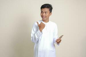 retrato do atraente ásia muçulmano homem dentro branco camisa segurando Móvel telefone com sorridente expressão em face enquanto apontando dedo para a lado. publicidade conceito. isolado imagem em cinzento fundo foto