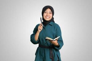 retrato do animado ásia muçulmano mulher com hijab escrevendo em Nota livro e apontando acima com caneta. publicidade conceito. isolado imagem em branco fundo foto
