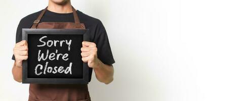 cortada retrato do ásia barista homem segurando uma quadro-negro segurando uma quadro-negro este diz Desculpe nós estão fechadas Boa para bandeira. isolado imagem em branco fundo. foto