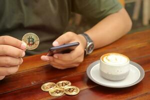 masculino mão segurando uma bitcoin enquanto usando telefone com uma copo do café em de madeira mesa dentro a cafeteria. criptomoeda negociação conceito foto