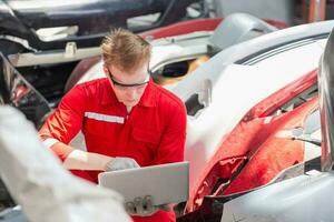 mecânico homem inspecionando carro partes estoque em computador portátil computador enquanto trabalhando dentro garagem armazém, jovem mecânico com computador portátil dentro auto reparar fazer compras foto