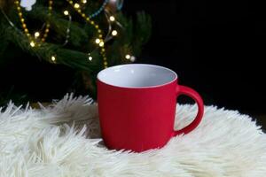 em branco vermelho caneca com Natal árvore em fundo, tapete chá ou café copo com Natal e Novo ano decoração, horizontal zombar acima com cerâmico caneca para quente bebidas, vazias presente impressão modelo foto