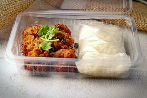 frito carne de porco colar e pegajoso arroz embrulhado colocada dentro uma plástico Leve embora caixa, em uma branco mesa. foto