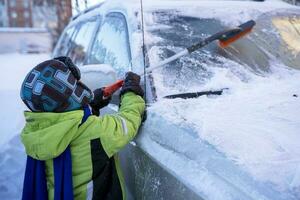 engraçado fofa caucasiano Garoto limpa a carro a partir de neve com uma escova e uma raspador foto