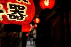 fechar-se Preto japonês Texto:% s em vermelho papel lanterna suspensão debaixo japonês restaurante beirais em noite Tempo com embaçado fundo. foto