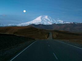 manhã panorama com enrolamento rodovia para montar Elbrus debaixo manhã céu com cheio lua. esvaziar estrada desaparecer para dentro a distância às crepúsculo. caminho com marcação perspectiva visualizar. kabardino-balkaria. foto