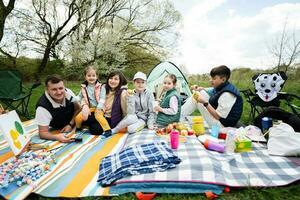 feliz jovem ampla família com quatro crianças tendo Diversão e desfrutando ao ar livre em piquenique cobertor às jardim Primavera parque, relaxamento. foto