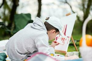 Garoto desfrutando ao ar livre em piquenique cobertor e pintura em tela de pintura às jardim Primavera parque, relaxamento. foto