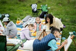 feliz jovem família, mãe e crianças tendo Diversão e desfrutando ao ar livre em piquenique cobertor pintura às jardim Primavera parque, relaxamento. foto