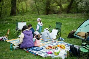 feliz jovem família, quatro crianças tendo Diversão e desfrutando ao ar livre em piquenique cobertor pintura às jardim Primavera parque, relaxamento. foto