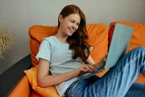 bonita mulher conversando em a laranja sofá com uma computador portátil tecnologias foto