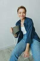 uma jovem mulher sentado dentro uma cadeira às casa sorridente com dentes com uma curto corte de cabelo dentro jeans e uma jeans camisa em uma branco fundo. menina natural poses com não filtros foto