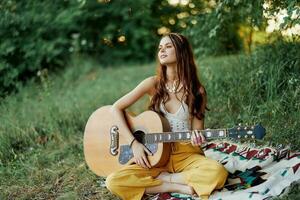 menina hippie mulher jogando guitarra dentro ecológico roupas sentado em a terra lado de fora dentro natureza dentro a outono assistindo a pôr do sol foto