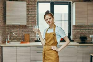 lindo mulher dentro a avental vidro do água dentro a cozinha tarefas domésticas casa vida foto