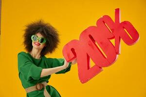 positivo jovem mulher verde vestir afro Penteado Sombrio óculos vinte por cento dentro mãos amarelo fundo inalterado foto