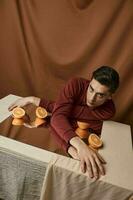 uma cara dentro uma camisa senta às uma mesa com uma espelho e laranja laranjas tecido fundo estúdio modelo morena quarto foto
