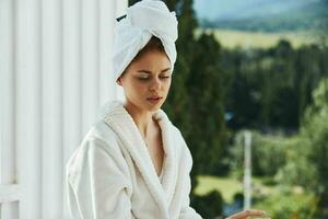 lindo mulher com uma toalha em meu cabeça dentro uma branco roupão de banho ficando em a sacada dentro uma hotel estilo de vida foto