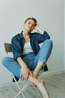 uma jovem mulher sentado dentro uma cadeira às casa sorridente com dentes com uma curto corte de cabelo dentro jeans e uma jeans camisa em uma branco fundo. menina natural poses com não filtros foto