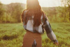 uma mulher corre através uma campo com dela costas para a Câmera em uma verão dia com dela cabelo grandes e vôo dentro a pôr do sol. a conceito do liberdade e harmonia com natureza foto