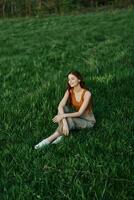 trabalhador autonomo mulher desfrutando a ao ar livre sentado dentro a parque em a verde Relva dentro casual roupas com grandes vermelho cabelo aceso de a brilhante verão Sol sem mosquitos foto