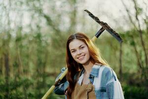 uma mulher sorridente belas e olhando às a Câmera, uma agricultor dentro trabalhos roupas e a avental trabalhando ao ar livre dentro natureza e segurando uma ancinho para reunir Relva e forragem para a animais dentro a jardim foto