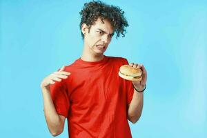 alegre cara com encaracolado cabelo dentro uma vermelho camiseta com uma Hamburger dentro dele mãos velozes Comida dieta foto