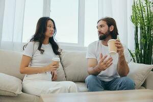 homem e mulher sentado às casa em a sofá dentro branco à moda Camisetas bebendo café Fora do caranguejo copos a partir de uma café fazer compras e tendo Diversão conversando sorrisos e riso às lar. masculino e fêmea amizade foto