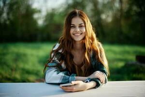 menina com telefone dentro mão sentado dentro natureza dentro a parque às a mesa sorridente belas e olhando às a Câmera com dela vermelho cabelo aceso de a pôr do sol luz solar do verão foto