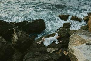 lindo mulher dentro uma isolado local em uma selvagem rochoso costa dentro uma branco vestir inalterado foto