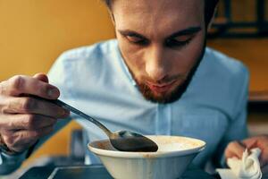 uma bonito homem com uma barba come borscht dentro uma prato às uma mesa dentro uma cafeteria e uma Assistir em dele mão foto