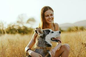 mulher e dela rouco cachorro alegremente passear através a Relva dentro a parque sorrir com dentes outono pôr do sol andar com bicho de estimação, viagem com uma cachorro amigo foto