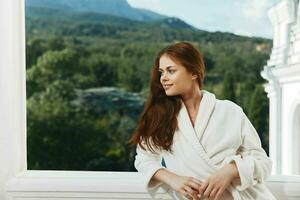 lindo mulher com grandes cabelo dentro uma branco roupão de banho ficando em a sacada dentro uma hotel montanha Visão foto