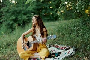 jovem fêmea hippie artista tocam guitarra e canta canções dentro ecológico roupas sentado em a terra lado de fora dentro natureza dentro a outono olhando Fora às a pôr do sol foto