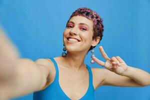 jovem Esportes mulher moda blogueiro leva uma cenário do ela mesma em a telefone dentro azul roupa de esporte sorridente e mostrando dela língua em uma azul monocromático fundo foto