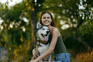 alegre mulher com uma rouco procriar cachorro sorrisos enquanto sentado dentro natureza em uma andar com uma cachorro em uma trela outono panorama em a fundo. estilo de vida dentro anda em com animais de estimação foto